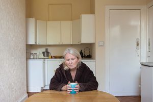 Photograph of woman at kitchen table clutching mug looking sad.